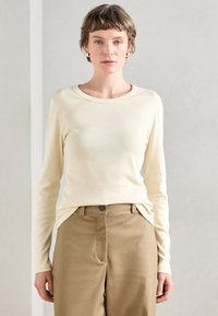 Πουκάμισο με μακριά μανίκια σε κρεμ χρώμα, με στρογγυλή λαιμόκοψη, φτιαγμένο από απαλή ύφασμα; συνδυασμένο με ανοιχτό καφέ, κομμένα παντελόνια που διαθέτουν πλαϊνές τσέπες.