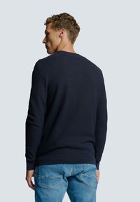 Marineblauwe gebreide trui met textuur, ronde halslijn, ribgebreide manchetten en zoom. Gekozen in combinatie met lichtblauwe jeans, achteraanzicht zichtbaar.