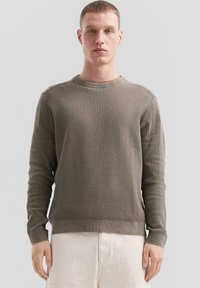 Olivgrüner Strickpullover mit strukturiertem Muster, runder Ausschnitt und gerippten Bündchen, kombiniert mit hellen Shorts.