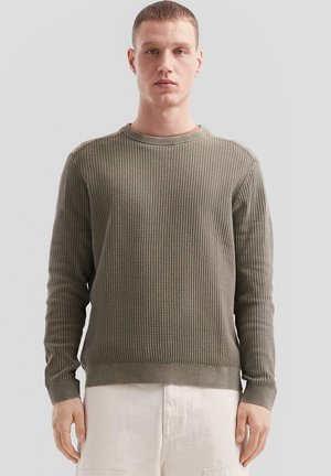 Olivgrüner Strickpullover mit strukturiertem Muster, runder Ausschnitt und gerippten Bündchen, kombiniert mit hellen Shorts.