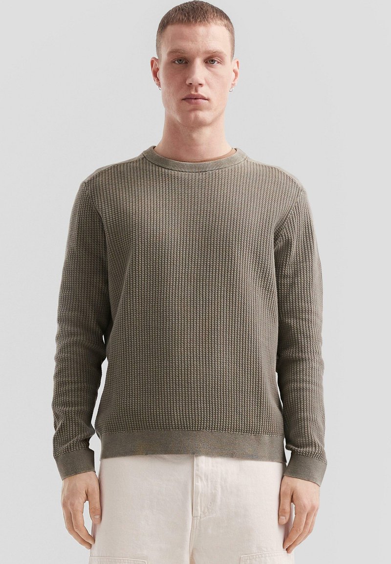Olivgrüner Strickpullover mit strukturiertem Muster, runder Ausschnitt und gerippten Bündchen, kombiniert mit hellen Shorts.