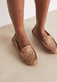 Des mocassins en toile marron présentent un motif de cercles superposés et un accent logo doré à l'avant. Couture douce et semelle flexible.