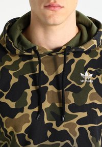 Mikina s kapucí v kamuflážovém vzoru v zelené a černé barvě. Obsahuje šňůrku na stažení a malé bílé logo Adidas na přední straně.