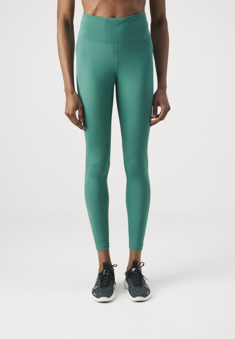 Persoon in hoge, turquoise leggings en zwarte sportschoenen, staand tegen een plain witte achtergrond.