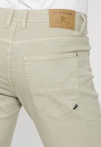 Pantalones de mezclilla beige con una etiqueta de cuero en la parte trasera de la cintura, que cuentan con un único bolsillo trasero y detalles de herrajes metálicos. Textura suave.