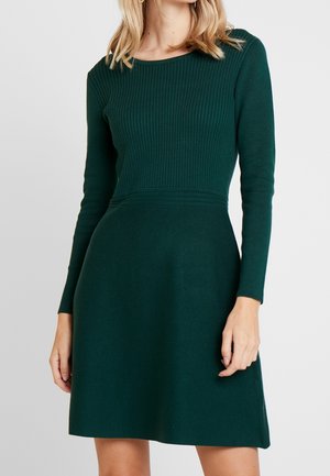 Kobieta ubrana w ciemnozieloną, długorękawową, prążkowaną dzianinową sukienkę z dopasowaną talią i rozkloszowanym dołem na jednolitym tle.