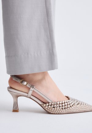 Mujer con zapato de tacón alto plateado con tira trasera adornado con grandes perlas, combinado con pantalones de pierna ancha en gris claro.