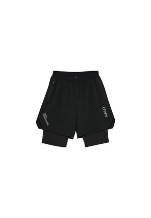 Jonge man staand, dragend een glanzend grijs jack met rits en capuchon, zwarte shorts met daaronder een legging, zwarte sokken en hardloopschoenen.
