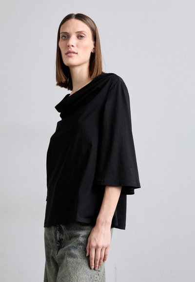 Black, oversized T-shirt with a draped neckline, wide short sleeves, and visible stitching details. Worn with light blue, textured jeans.