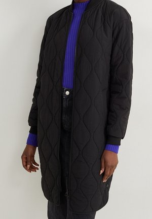 Long manteau noir matelassé porté sur un pull violet côtelé et un pantalon noir, présenté sur une personne debout avec les bras détendus.