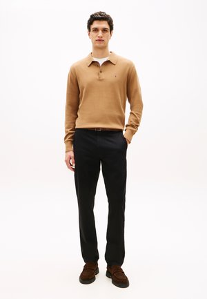 Uomo con polo marrone cammello a manica lunga, pantaloni neri, mocassini marroni, in piedi con una mano in tasca su sfondo bianco.