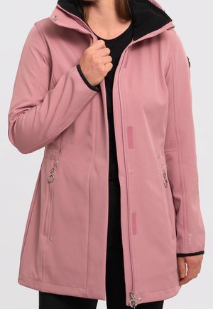 Vrouw draagt een zachte roze jas met rits en capuchon en ritszakken aan de zijkant, houdt de ritssluiting bij haar borst vast over een zwart shirt.