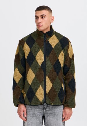 Mann trägt eine Fleecejacke mit Reißverschluss mit großem Rautenmuster in Olivgrün, Braun, Beige und Schwarz, steht vor einem schlichten Hintergrund.