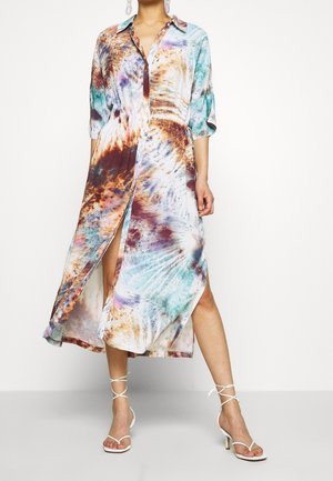 Kvinde iført en farverig, langærmet, knap-ned tie-dye midi kjole med slidser foran og hvide hælede sandaler med stropper.