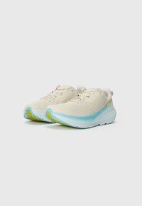 Chaussures de sport beiges avec tige en mesh, semelle bleu clair et accents verts. Présente un design texturé et un système de fermeture traditionnel à lacets.