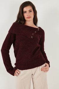 Naine, kes kannab burgundia värvi kootud kampsunit asümmeetrilise nööbitava kraega ja heledaid beeže pükse, seisab ühevärvilise tausta ees.