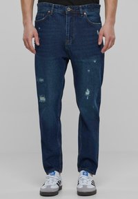 Modri denim kavbojke z poškodovanimi detajli, ravnega kroja, petimi žepi in kovinskimi dodatki. V kombinaciji z belimi supergami.