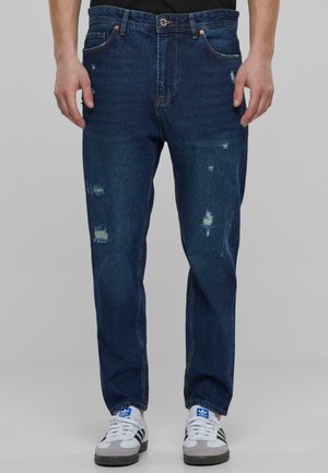 Jean en denim bleu avec des détails usés, coupe droite, cinq poches et quincaillerie métallique. Associés à des baskets blanches.