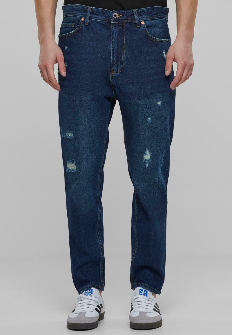 Modri denim kavbojke z poškodovanimi detajli, ravnega kroja, petimi žepi in kovinskimi dodatki. V kombinaciji z belimi supergami.
