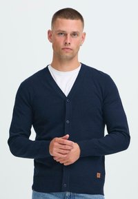 Fiatal férfi rövid hajjal, sötétkék gombos kardigánt visel egy fehér ing felett, és egy sima világos háttér előtt áll.