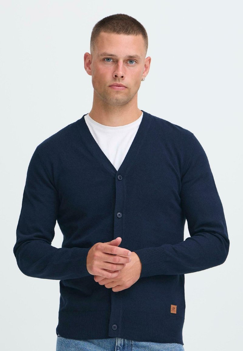 Fiatal férfi rövid hajjal, sötétkék gombos kardigánt visel egy fehér ing felett, és egy sima világos háttér előtt áll.