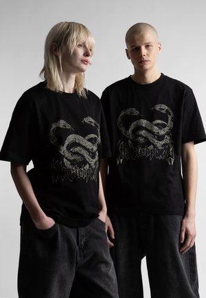 Dwoje młodych dorosłych w czarnych koszulkach z grafiką dwóch splecionych węży i ciemnych dżinsach, stojących na tle gładkiego tła.