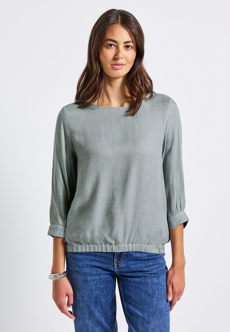 Blusa verde chiaro, ampia e con maniche a tre quarti, con orlo arricciato. Tessuto morbido e testurizzato con scollo quadrato.