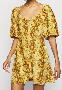 Kortärmad, knälång klänning i gult med ett ormskinsmönster. Har en fyrkantig halsringning och rynkade detaljer i mitten.