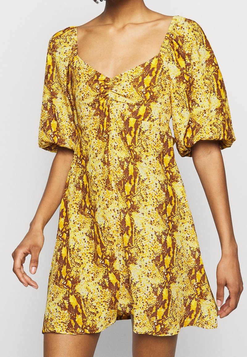Kortärmad, knälång klänning i gult med ett ormskinsmönster. Har en fyrkantig halsringning och rynkade detaljer i mitten.