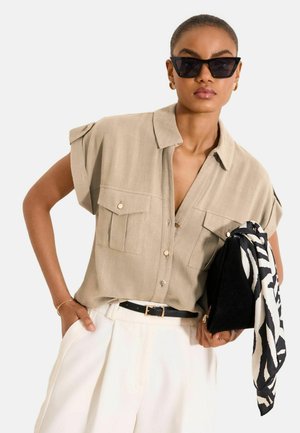 Naine, kellel on seljas beež lühikeste varrukatega nööpidega särk, valged kõrge vöökohaga püksid musta vööga, mustad kassisilma päikeseprillid, hoides musta klambriga zebra mustriga salli.