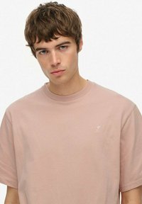 Ung mand med kort brunt hår og blå øjne iført en ensfarvet lyserød t-shirt med rund hals mod en hvid baggrund.