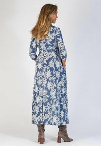 Maxiklänning i blå denim med vitt blommönster; har långa ärmar och fickor. Kombinerad med grå mockakängor till knäna.