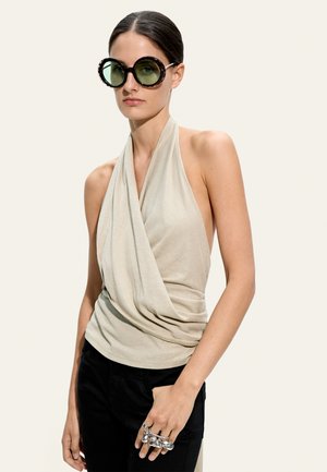 Kobieta z zaczesanymi do tyłu włosami, nosząca duże okrągłe okulary przeciwsłoneczne, beżowy plisowany top na szyję, czarne spodnie i srebrne pierścionki na kostkach palców.