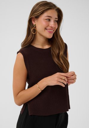 Jonge vrouw die lacht, draagt een mouwloos donkerbruin gebreid topje, gouden oorringen, ringen en een armband tegen een neutrale achtergrond.