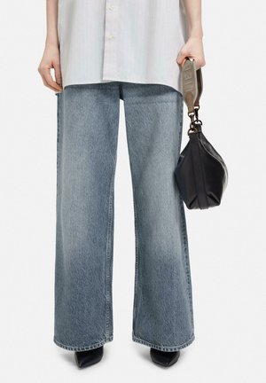 Osoba nosząca luźne niebieskie dżinsy i czarne, spiczaste buty, trzymająca czarną torebkę z szerokim beżowym paskiem.