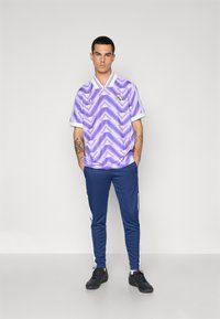 Camisa de fútbol de patrón morado con cuello y puños blancos, combinada con pantalones deportivos azul marino con franjas laterales blancas. Zapatillas negras.