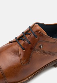 Chaussure en cuir marron avec des lacets noirs, bout rond et coutures décoratives. Présente une surface texturée et un accent en métal sur le côté.