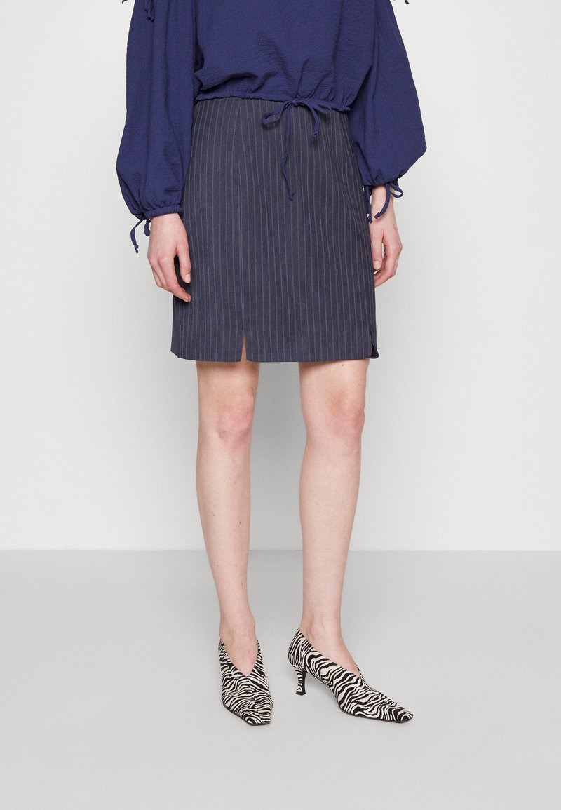 Jupe plissée bleu marine avec des rayures verticales, assortie à des escarpins à bout pointu en imprimé zèbre noir et blanc. Blouse texturée avec des détails à cordon.