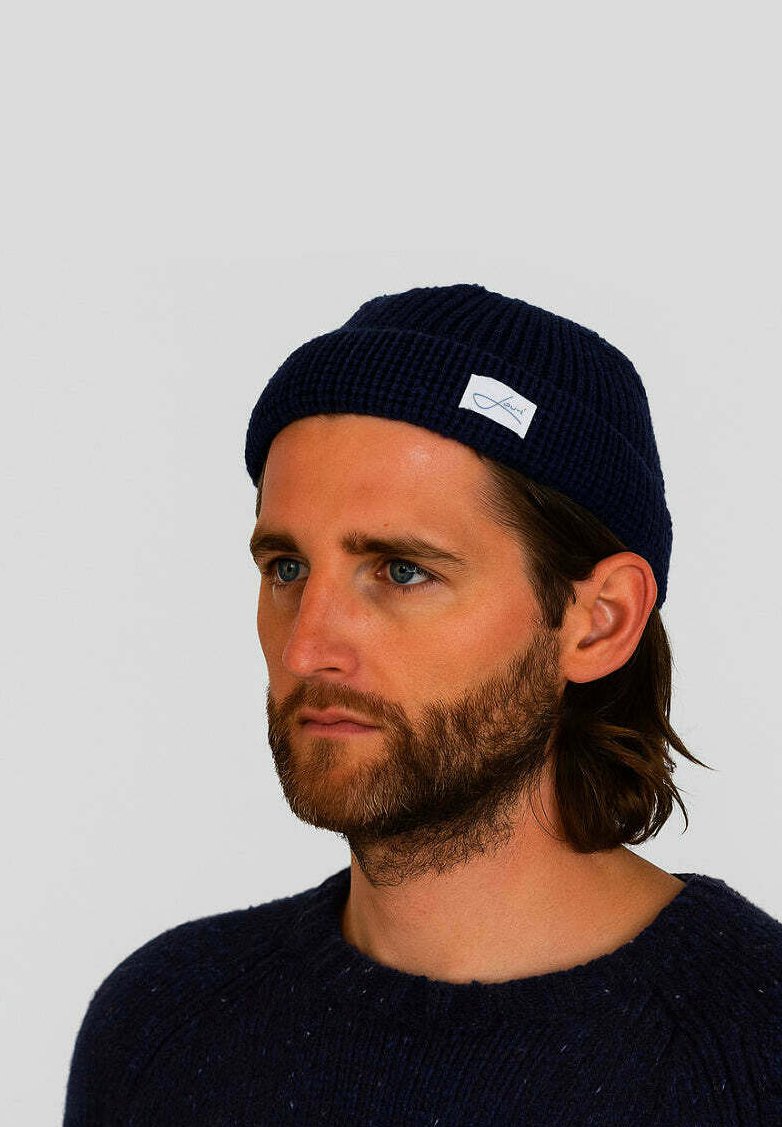 Navy knitted beanie with ribbed texture and a small white label accent. Worn with a dark sweater, features a snug fit.