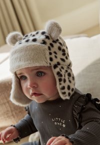 Nažehlená béžová čepice s černými leopardími skvrnami, klapkami na uši a zaoblenými ušima. Dítě oblečené v šedém dlouhém rukávu s volánkovými detaily.