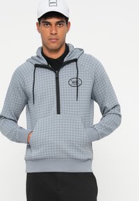 Grå zip-up hettegenser med små rutemønstre, kengurulomme, svarte detaljer og ribbestrikkede mansjetter. Bærer logo på brystet.