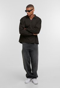 Black long-sleeve polo with a V-neck, paired with loose gray jeans and white sneakers. The model wears black sunglasses, standing with arms crossed.