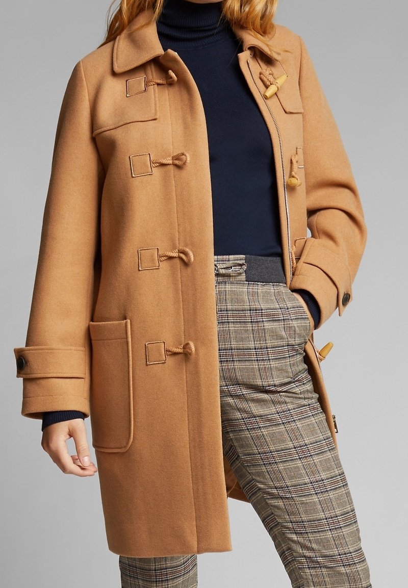 Femme portant un manteau camel à brandebourgs, un col roulé bleu marine et un pantalon à carreaux taille haute, les mains partiellement dans les poches.