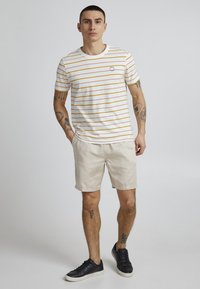 White short-sleeve shirt with yellow stripes, featuring a small logo, paired with beige shorts and black sneakers. Smooth fabric texture.