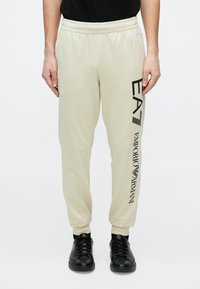 Smėlio spalvos laisvalaikio kelnės su elastinga juosmens juosta. Šone yra juodas užrašas „EA7 EMPORIO ARMANI“. Lygus, lengvas audinys.