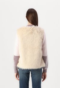 Gilet en fausse fourrure beige avec une texture shaggy, encolure arrondie et sans fermetures. Porté par-dessus un pull multicolore à rayures et associé à un jean bleu.