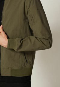 Olivegrüner Bomberjacke aus leichtem Material, mit gerippten Bündchen und Saum, seitlichen Taschen und einem dezenten Logo-Detail am Ärmel.