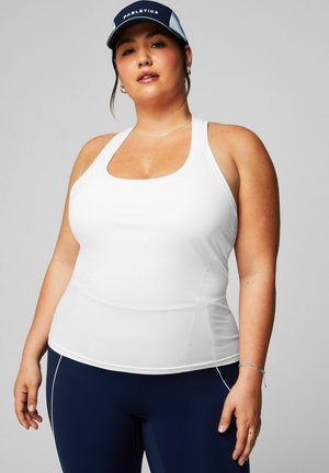 Frau trägt ein weißes Tanktop, marineblaue Leggings mit weißen Paspeln, eine marineblaue und weiße Kappe sowie silbernen Schmuck, steht vor einem grauen Hintergrund.