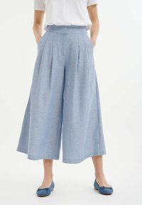 Lichtblauwe wijde broek met hoge taille, plooien en zijzakken. Gemaakt van een gestructureerde stof. Gepaard met blauwe platte schoenen.