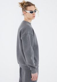 Grauer Sweatshirt mit Rundhalsausschnitt, ribbed Bündchen und lockerer Passform. Das Model trägt außerdem weiße futuristische Sonnenbrillen.